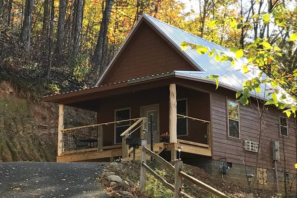 Small wooden cabin with metal roof and front porch, surrounded by green hills and larger cabin in background