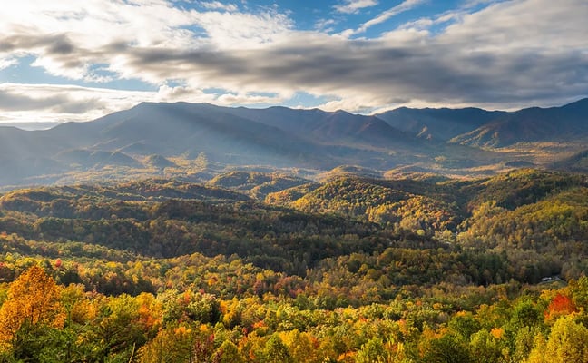 Autumn panorama of the Great Smoky Mountains, with layers of forested ridges in gold, orange and green under a partly cloudy sky.